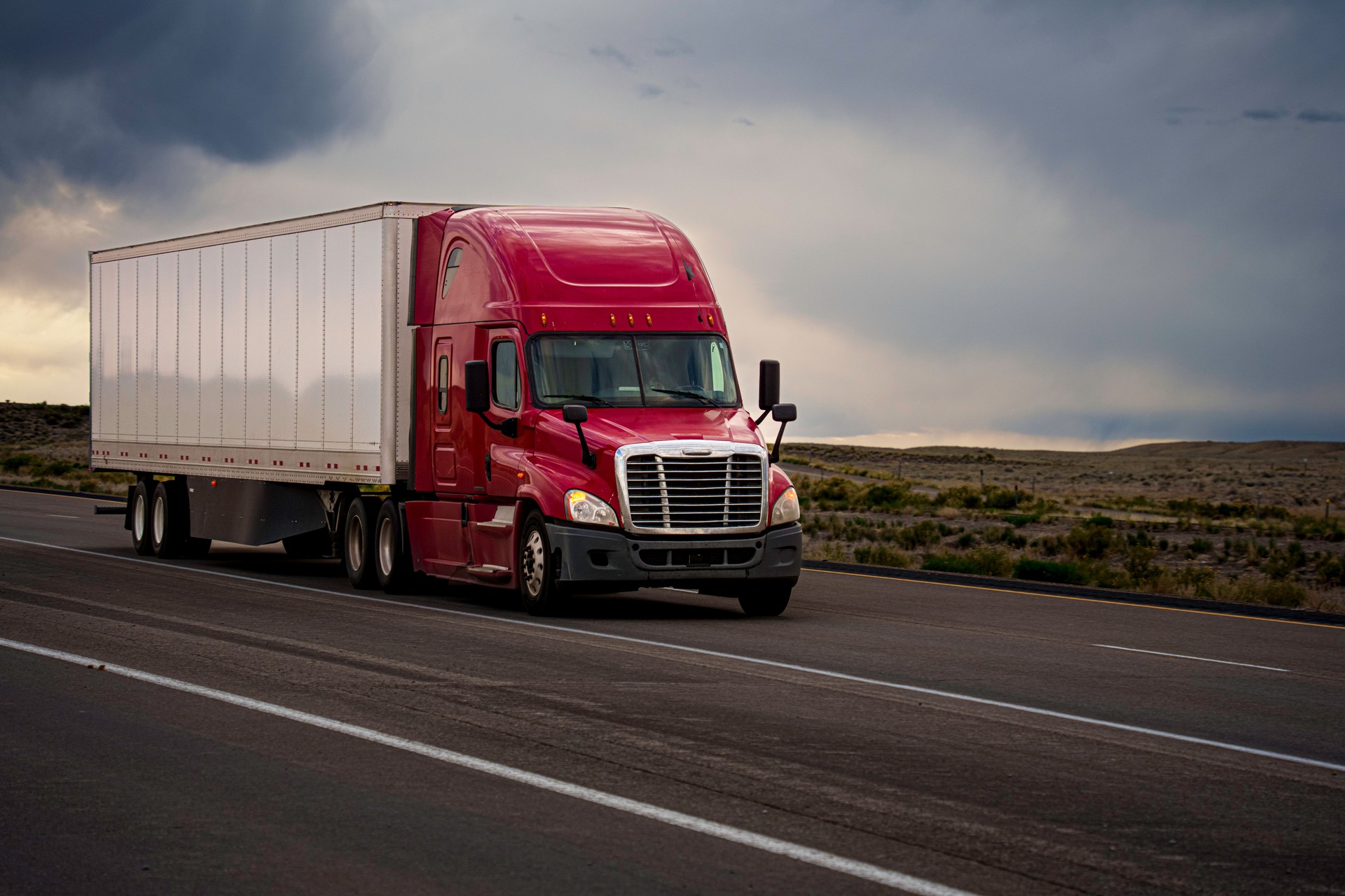 Low Angle Red Semi-Trailer Long Haul Tractor Truck Speeding Down a Four Lane Highway in Eastern Utah Desert